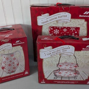 Puede incluir: Tres cajas rojas con diseños de copos de nieve blancos y la palabra "Benevolence" impresa en ellas. Las cajas contienen una jarra de servir de cerámica, una bandeja de servir cuadrada y un servidor de vacaciones de dos niveles con una bandeja de metal.