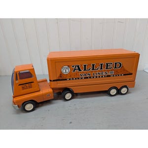 May include: An orange toy semi-truck with a trailer featuring the text Allied Van Lines, Inc. World's Largest Mover. The truck has white wheels, a cab with a blue windshield, and black lettering on the side of the trailer and the cab door.