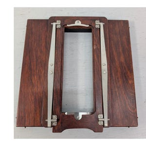 May include: A vintage wooden camera accessory with a rectangular frame and two hinged side panels. The dark brown wood is accented by silver-colored metal hinges and supports. The item appears to be a part of a larger photographic setup.