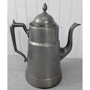 May include: A tall, metallic gray pewter coffee pot with a cylindrical body, a hinged lid topped with a pointed finial, a curved spout, and an angular handle. The surface shows a weathered, matte finish with visible signs of age and use.