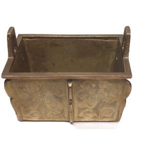 May include: A rectangular brass container with floral engravings. The container has two handles and is likely used for decorative purposes. The sides feature detailed floral patterns, adding an antique aesthetic.