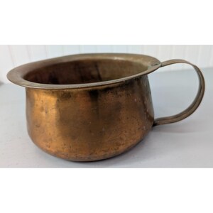 May include: A vintage copper-toned chamber pot with a rounded body and a handle. The pot has a wide, slightly flared rim and shows signs of age and use, with a patinaed finish. The handle is a simple, curved design.