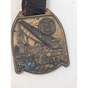 May include: A brass-toned metal fob featuring a relief image of a crane with the text "THEW CENTER DRIVE" and "LORAIN" embossed on the surface. The piece has a dark, weathered patina and is attached to a worn, dark brown leather strap at the top.