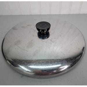May include: A stainless steel pot lid with a black handle. The lid is round and has a shiny, reflective surface. The black handle is circular and sits in the center of the lid. The lid is designed for cookware.