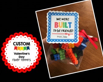 May include: A blue and white printable Valentine's Day card with the text "We were BUILT to be friends!" and "From, Jake". The card is decorated with white polka dots. A bag of colorful plastic building blocks is in the background.