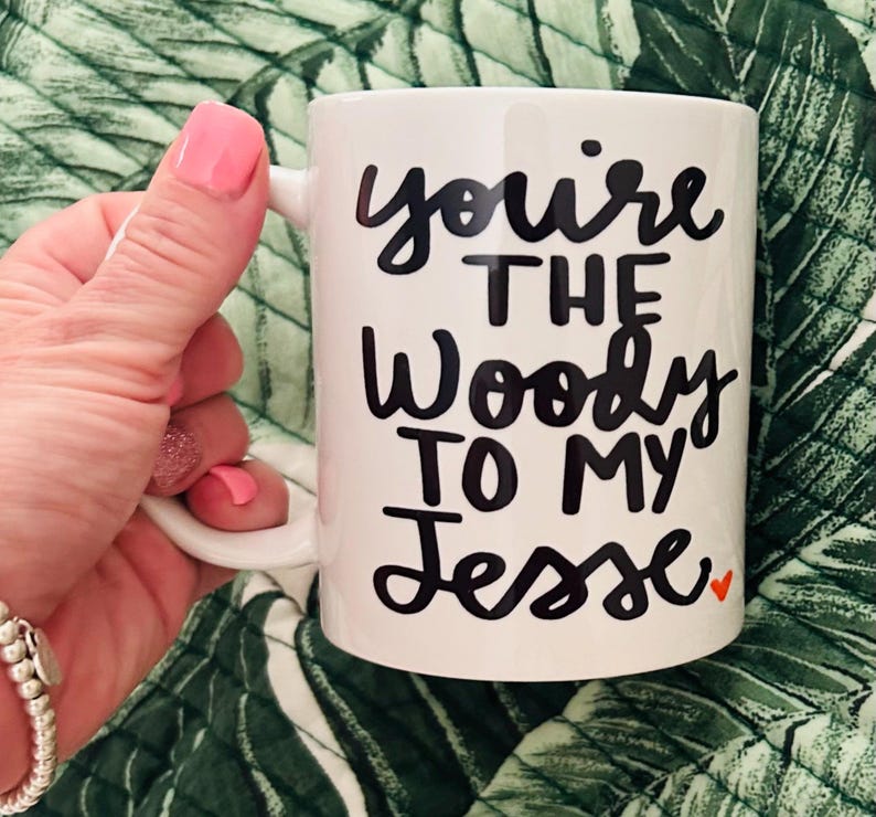 May include: A white ceramic mug featuring black text that reads "you're THE Woody TO MY Jesse" with a small orange heart at the end of the final word. The mug has a curved handle on the left side and a smooth, glossy finish throughout the exterior.