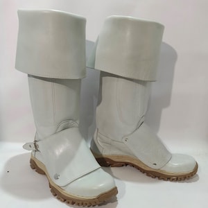 Puede incluir: Un par de botas de cuero blanco con un puño abatible y una correa con hebilla. Las botas tienen una suela de goma marrón con un patrón de banda de rodadura.