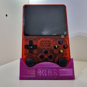 May include: A transparent red handheld gaming console with black buttons and a black screen, resting on a purple stand. The stand has the text "R36S" on it. The console features a directional pad, action buttons, and start/select buttons.