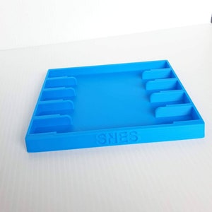 May include: A blue plastic game cartridge storage case with slots for multiple cartridges. The case has the word "SNES" printed on the front.