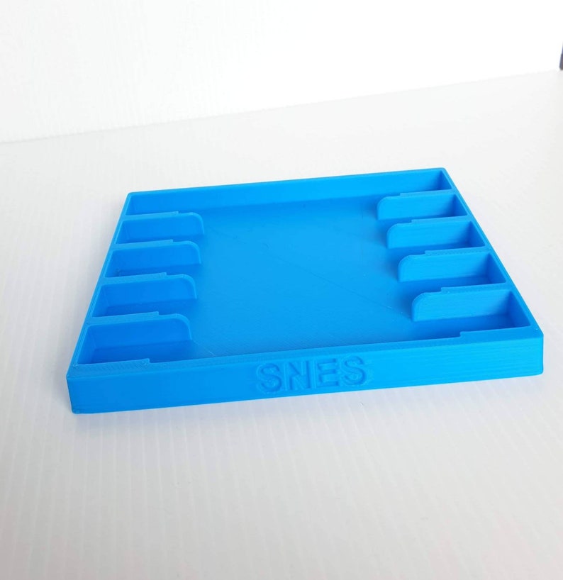 May include: A blue plastic game cartridge storage case with slots for multiple cartridges. The case has the word "SNES" printed on the front.