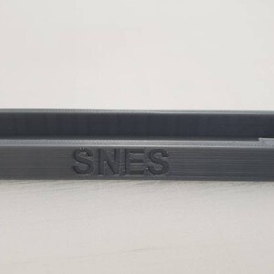 May include: A gray plastic storage box with the word "SNES" printed on the front.