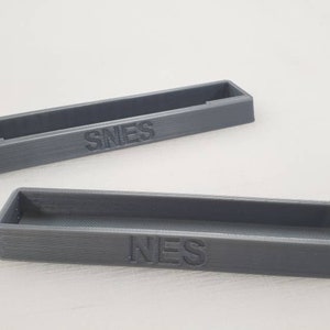 May include: Two gray plastic storage containers with the words "NES" and "SNES" printed on them.