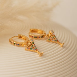 May include: A pair of gold hoop earrings with a dangling martini glass charm. The glass is decorated with colorful gemstones.