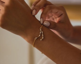 May include: A gold-toned chain bracelet featuring a constellation design composed of small, clear, round-cut stones. The delicate metal chain connects to the central star-shaped arrangement, which is secured with thin gold-colored metal links.