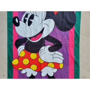 Pu&ograve; includere: Un asciugamano da spiaggia colorato con un'immagine di cartone animato di Minnie Mouse che indossa una gonna a pois rossi e gialli. Lo sfondo &egrave; viola con strisce rosa e verdi.
