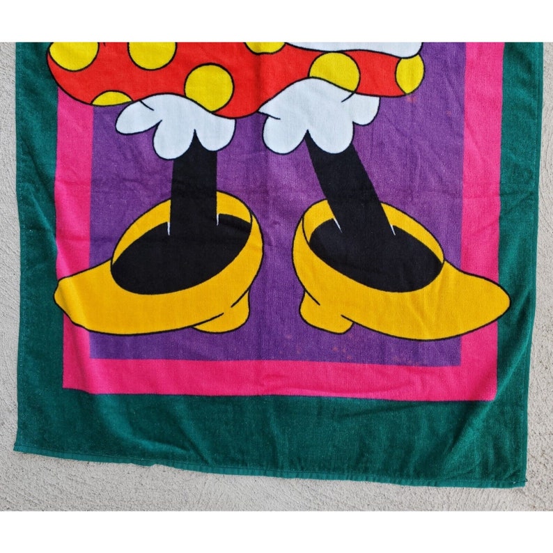Pu&ograve; includere: Un asciugamano da spiaggia verde, rosa e viola con un'immagine di cartone animato di Minnie Mouse che indossa scarpe gialle e un vestito a pois rossi e bianchi.