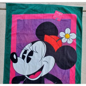 Pu&ograve; includere: Un asciugamano da spiaggia verde, rosa e viola con un'immagine di cartone animato di Minnie Mouse. Minnie Mouse indossa un fiocco rosso e ha un fiore bianco sulla testa. L'asciugamano ha il testo "Mickey's" in un riquadro rosa.