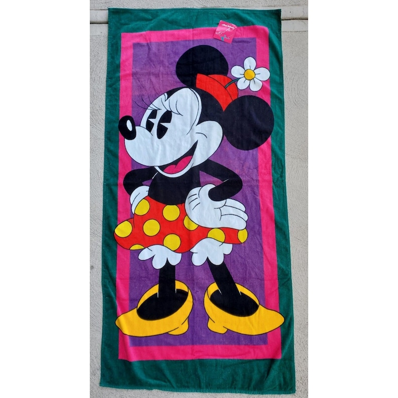 Pu&ograve; includere: Un asciugamano da spiaggia verde con un bordo rosa e un centro viola. Un'immagine di cartone animato di Minnie Mouse &egrave; al centro dell'asciugamano. Minnie Mouse indossa un vestito a pois rossi e bianchi e un fiocco rosso. Ha un fiore giallo nei capelli.