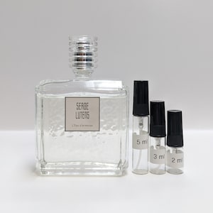May include: A clear glass bottle of Serge Lutens L'Eau d'Armonie perfume with a silver cap. Three smaller sample bottles are next to the larger bottle, labeled 5 ml, 3 ml, and 2 ml.