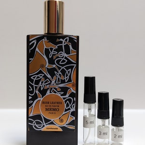 May include: Three glass perfume sample vials with black caps and white labels. The labels read "5 ml", "3 ml", and "2 ml". The vials are in front of a larger bottle of perfume with a black and gold label that reads "Irish Leather Eau de Parfum Memo Paris Cuirs Nomades".