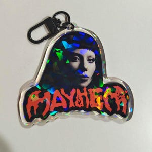 May include: A clear acrylic keychain featuring a woman's portrait with a holographic effect. The word "MAYHEM" is printed in red and orange. The keychain has a black clasp.