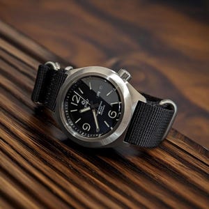 Può includere: Un orologio da polso in acciaio inossidabile con quadrante nero e cinturino in nylon nero. L'orologio ha numeri e lancette bianche, con i numeri 12, 6 e 9 ben visibili. L'orologio è impermeabile e automatico.