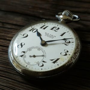 May include: A silver-toned Seiko Precision 15 Jewels pocket watch with a white dial, black Arabic numerals, and a sub-dial for seconds. The watch features a winding crown at the top and black hands, with text reading Seiko Precision 15 Jewels.