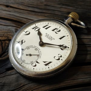 May include: A vintage Seiko pocket watch with a white face, black numerals, and a second dial. The watch has a silver case and a gold-colored crown. The watch face reads "SEIKO PRECISION 15 JEWELS".