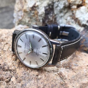 May include: Vintage Seiko Sportsman watch with a silver dial, silver hands, and a black leather strap. The watch face has the text "WATER PROOF" and "17 Jewels". The watch is set on a rocky surface.
