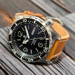 May include: A stainless steel Level7 dive watch with a black dial, white numerals, and a date display. The watch has a rotating bezel and a tan leather strap. The watch is water resistant to 20 bar.