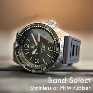 Op de afbeelding: Een roestvrijstalen horloge met een donkergroene wijzerplaat, grote witte cijfers en een grijze rubberen band. De wijzerplaat geeft de dag van de week en de tijd weer. Waterbestendig tot 20 bar.