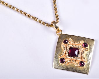 May include: A gold necklace with a square pendant featuring a red gemstone surrounded by smaller gemstones. The pendant has a textured surface and a rope chain.