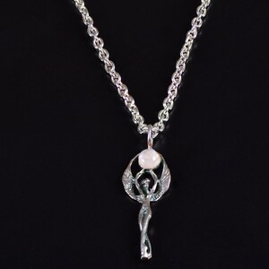 May include: A silver-toned pendant necklace featuring a stylized figure with outstretched wings, holding a round, white stone. The pendant hangs from a silver chain against a black background. The necklace is a piece of jewelry.