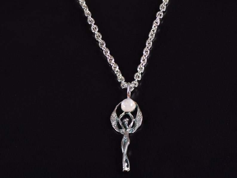 May include: A silver-toned pendant necklace featuring a stylized figure with outstretched wings, holding a round, white stone. The pendant hangs from a silver chain against a black background. The necklace is a piece of jewelry.