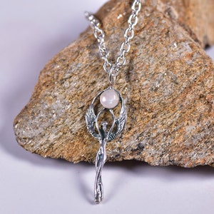 May include: A silver-toned pendant necklace featuring a stylized angel design. The pendant has a central figure with outstretched wings, a round, light-colored stone, and a twisted lower portion. The necklace is displayed on a textured, brown rock.