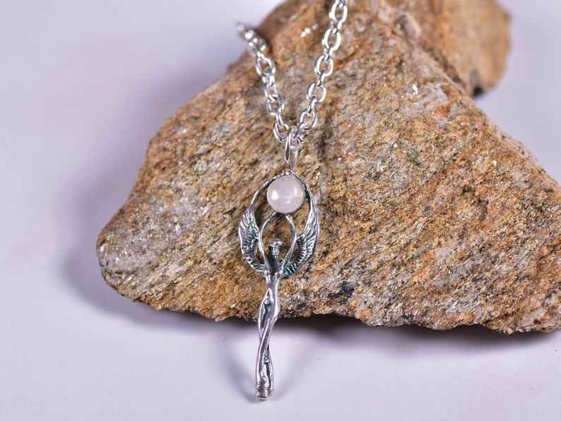 May include: A silver-toned pendant necklace featuring a stylized angel design. The pendant has a central figure with outstretched wings, a round, light-colored stone, and a twisted lower portion. The necklace is displayed on a textured, brown rock.