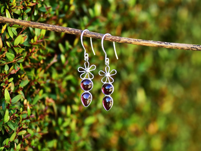 May include: A pair of silver dangle earrings with three red gemstone beads and a flower design at the top.