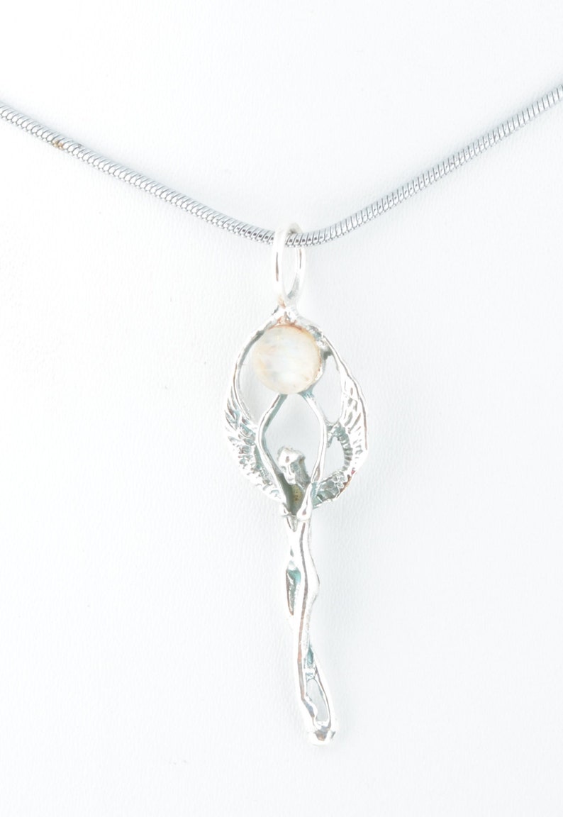 May include: A silver pendant necklace featuring a stylized figure holding a round, off-white stone. The pendant has an angel-like design with outstretched wings. The necklace chain is a thin, silver snake chain.