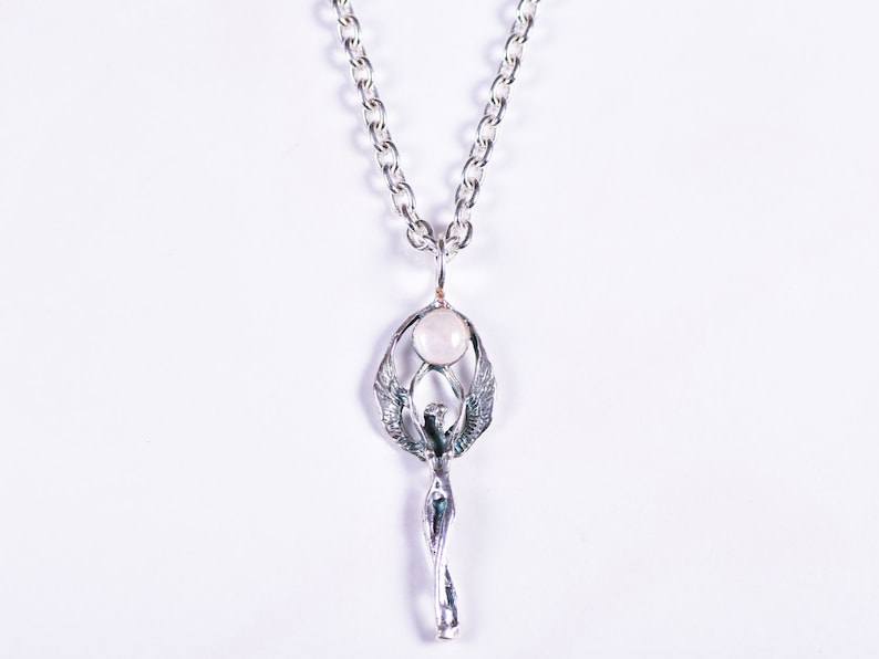 May include: A silver-toned pendant necklace featuring a stylized figure with outstretched wings, holding a round, pale pink stone. The pendant hangs from a silver chain. The design is intricate and detailed, with a focus on the figure's form.