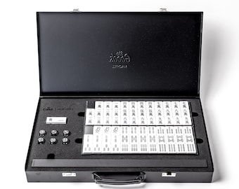 Puede incluir: Una maleta de transporte negra con un cierre y un asa plateados. La maleta está abierta, revelando un juego de fichas de mahjong con caracteres chinos y números. Las fichas están dispuestas en filas y columnas, y también hay cuatro pequeños dados blancos.