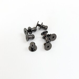 May include: A group of six black metal push button fasteners with a screw-on design. The fasteners have a round head and a small, round knob.