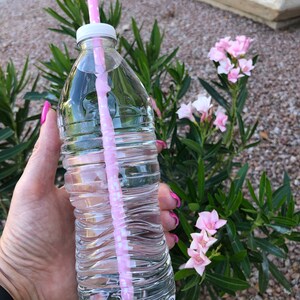 May include: A clear plastic water bottle with a pink and white striped straw. The bottle is filled with water and has a white lid. The bottle is being held by a hand with pink nail polish.