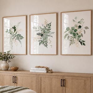 May include: A bedroom scene featuring a bed with a cream-colored comforter and a green throw blanket. Three framed botanical prints hang above a wooden cabinet. A lamp sits on a nightstand, and a window with sheer curtains is visible.
