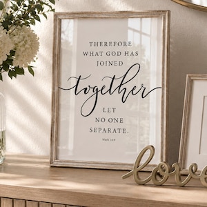 Può includere: Una stampa artistica da parete incorniciata verticalmente con il testo nero Therefore what God has joined together let no one separate Mark 10:9. La stampa è esposta in una cornice di legno chiaro accanto a una piccola illustrazione di un albero incorniciata e a una scritta love in metallo.