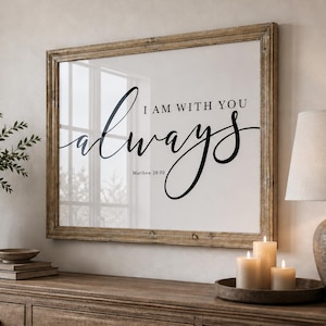 Può includere: Una stampa incorniciata con le parole "I AM WITH YOU always" in corsivo nero. La stampa è esposta sopra un comò in legno, accanto a una lampada, candele e un vaso con piante. L'estetica generale è calda e accogliente.
