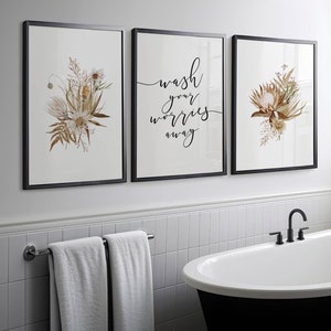 May include: Three framed prints with black frames. The centre print has the text "Wash your worries away" in black script. The left and right prints feature dried floral arrangements with white and brown flowers.