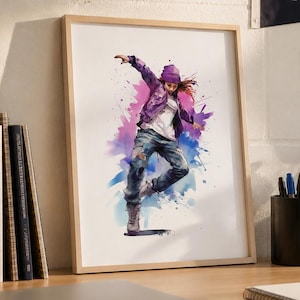 May include: A framed watercolor print of a dancer in motion, wearing a purple jacket, ripped jeans, and a beanie. The artwork features vibrant splashes of pink, blue, and purple. The print is displayed on a wooden desk with books, a laptop, and a water bottle.