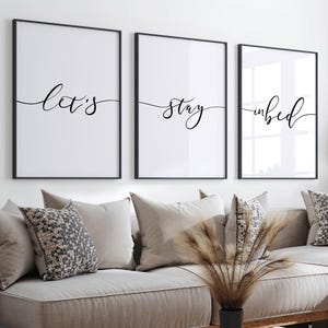 May include: Three black framed prints with white backgrounds and black calligraphy. The prints read "Let's", "stay", and "in bed".