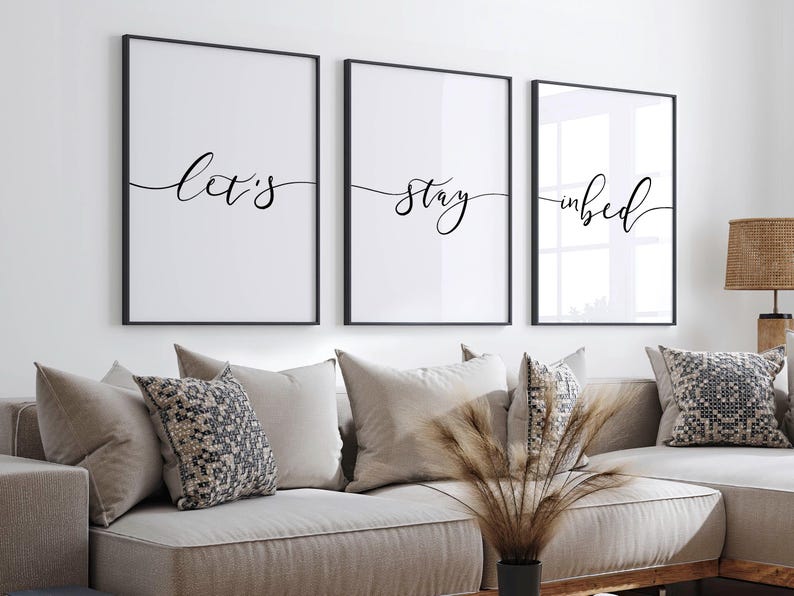 May include: Three black framed prints with white backgrounds and black calligraphy. The prints read "Let's", "stay", and "in bed".