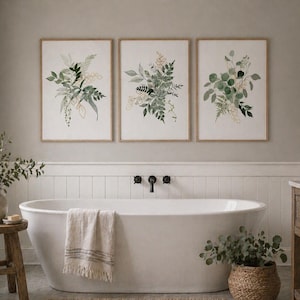 Puede incluir: Tres impresiones art&iacute;sticas bot&aacute;nicas enmarcadas con ilustraciones en acuarela de follaje verde y dorado. La obra de arte se exhibe sobre una ba&ntilde;era blanca en un ba&ntilde;o. La habitaci&oacute;n tiene una paleta de colores neutros con detalles en madera natural.
